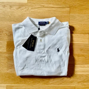 Ralph lauren pike - Klassisk vit pikétröja från Polo Ralph Lauren med broderad logga på bröstet. Tröjan har krage, knappar vid halsen och är kortärmad. Perfekt för en stilren och tidlös look. Helt ny o har etiketten på. Storlek S. 