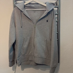 Grå hoodie med dragkedja från Polo Ralph Lauren - Säljer en klassisk grå hoodie från Polo Ralph Lauren med dragkedja och huva. Tröjan har två fickor framtill och en broderad svart logga på bröstet. Perfekt för en avslappnad stil.