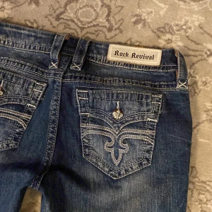 Rock Revival jeans - Säljer ett par blå Rock Revival jeans med snygga broderade detaljer på bakfickorna och slitningar framtill. Jeansen har raka ben och klassisk femficksdesign. Märkespatch i midjan och dekorativa sömmar ger extra edge.