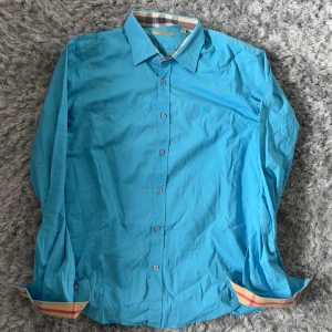 Blå skjorta från Burberry - Säljer en snygg blå skjorta från Burberry med klassiskt rutigt mönster på insidan av kragen och ärmsluten. Skjortan har långa ärmar och knäppning framtill. Perfekt för dig som gillar stilrena plagg med en twist.