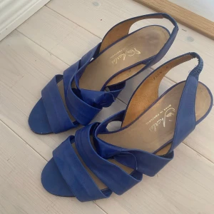 Blåa sandaler med kilklack från Acarles - Snygga blå sandaler från Acarles med kilklack och öppen tå. Skorna har breda remmar som korsar över foten och en elastisk rem runt hälen för extra komfort. Perfekta för varmare dagar och ger en stilren look.