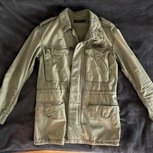 Polo Ralph Lauren jacka - Säljer en grön field jacka från Polo Ralph Lauren med US-RL brodyr på bröstet. Jackan har flera stora fickor framtill, klassisk krage och knapp- samt dragkedjestängning. Perfekt för dig som gillar en stilren och funktionell look.