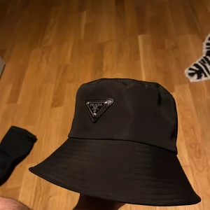 Svart bucket hat från Prada - Stilren svart bucket hat från Prada med klassisk logga framtill. Hatten har en mjuk och följsam form och är tillverkad i ett slätt material som ger en exklusiv känsla. Perfekt accessoar för att lyfta din outfit.