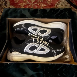 Dior b30 - Säljer ett par svarta Dior sneakers med stora reflekterande CD-loggor på sidorna och vita snören. Skorna har en vit, tjock sula och sportig design. Perfekta för dig som vill sticka ut med exklusiva och trendiga sneakers. Aldrig använd eftersom att skorna är för små