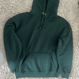 Mörkgrön hoodie med ficka - Säljer en mörkgrön hoodie med huva och dragsko. Tröjan har en stor magficka och ribbade muddar vid ärmslut och nederkant. Perfekt för en avslappnad stil.