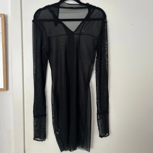 Svart meshklänning med lång ärm - Säljer en svart, transparent meshklänning med lång ärm och v-ringad hals. Klänningen är tight och har en minimalistisk design som passar perfekt för lager-på-lager eller som statement-plagg.