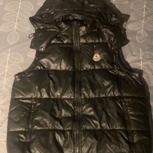 Svart dunväst från Moncler - Säljer en svart, glansig dunväst från Moncler med huva och dragkedja framtill. Västen har Moncler-logga på bröstet och två fickor med dragkedja. Perfekt för lager-på-lager och streetwear. Priset kan diskuteras skriv för minsta fundering
