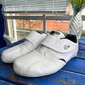 Vita sneakers från Lacoste med kardborreband - Säljer ett par vita sneakers från Lacoste med kardborreband och svart detalj vid hälen. Skorna har rund tå, platt sula och är tillverkade i syntetmaterial. Perfekta för dig som gillar sportig stil och enkel design.
