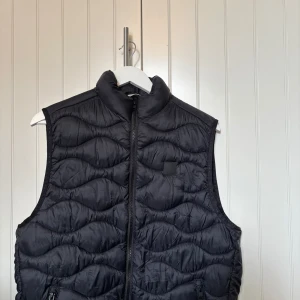 Svart dunväst från Jack & Jones - Säljer en svart dunväst från Jack & Jones i storlek M. Västen har dragkedja framtill och två fickor med dragkedja. Quiltad design med vågigt mönster och hög krage. Små spår på användning.