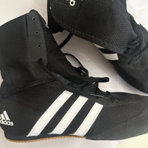 Svarta Adidas boxningsskor med vita ränder - Säljer ett par svarta Adidas boxningsskor med klassiska vita ränder på sidorna och gummisula. Skorna har hög modell, snörning hela vägen upp och är tillverkade i syntetmaterial. Perfekta för träning inom boxning eller kampsport.