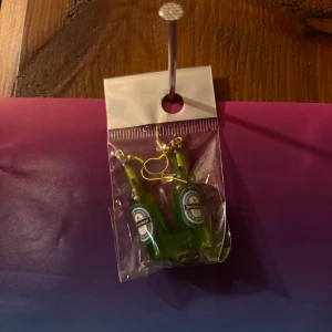 Örhängen med gröna flaskor - Säljer ett par unika örhängen formade som gröna flaskor med etikett. Perfekt accessoar för dig som vill sticka ut och addera en rolig detalj till din stil.