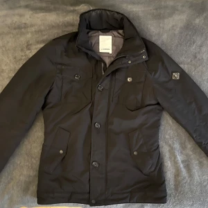 J.Lindeberg jacka - Snygg svart jacka från J.Lindeberg med hög krage och knäppning framtill. Jackan har fyra fickor med lock och knappar samt en diskret logotyp på ärmen. Perfekt för kyligare dagar.