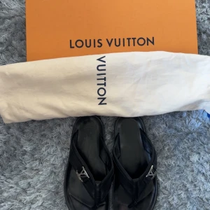 Svarta sandaler från Louis Vuitton loafers skor flippflopps - Säljer ett par svarta sandaler från Louis Vuitton i skinn med silverfärgad LV-logga på remmen. Klassisk och stilren design, perfekt för sommaren. Levereras med originalpåse och låda.