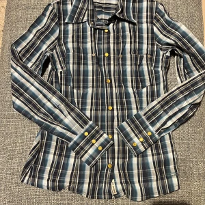 Blå och vit rutig skjorta från Peak Performance - Snygg rutig skjorta i blått, vitt och beige från Peak Performance. Skjortan har långa ärmar, klassisk krage och gula knappar framtill samt vid ärmsluten. Perfekt för dig som gillar en stilren och tidlös look.