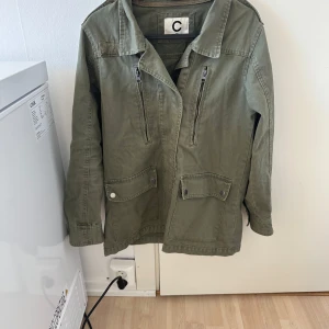 Olivgrön jacka med dragkedjor från Cubus - Säljer en olivgrön jacka från Cubus med två bröstfickor med dragkedja och två stora fickor med lock och knapp framtill. Jackan har klassisk krage och lång ärm, perfekt för lager på lager under höst och vår.