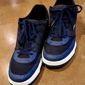 Blå sneakers från Lacoste - Säljer ett par snygga blå sneakers från Lacoste med vita och mörkblå detaljer. Skorna har snörning, rund tå och platt sula. Perfekta för dig som gillar stilrena och sportiga skor med klassisk Lacoste-logga på sidan.