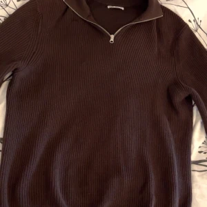 Brun ribbstickad tröja med dragkedja från Zara - Mörkbrun ribbstickad tröja från Zara med halv dragkedja vid halsen. Tröjan har lång ärm och hög krage, perfekt för kyligare dagar. Enkel och stilren design som passar till mycket.