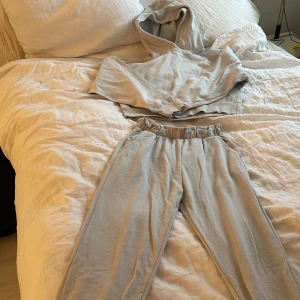 Ljusgrå mjukisbyxor med resår - Säljer ett par ljusgrå mjukisbyxor som kommer med en matchande tröja med resår i midjan och vid bensluten. Byxorna har en avslappnad passform och är tillverkade i mjukt bomullsmaterial, perfekta för en chill vardagslook.