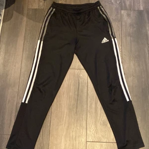 Svarta träningsbyxor från Adidas med vita ränder - Säljer ett par svarta Adidas träningsbyxor i storlek 11-12 år (152 cm). Byxorna har klassiska vita ränder längs sidorna och Adidas-logga på låret. Tillverkade i Primegreen-material och har resår i midjan. Perfekt för träning eller en komplett look. En inte så stor defekt så gör ingen påverkan på själva användningen. För bara 80kr så får du de bästa adidas byxorna!