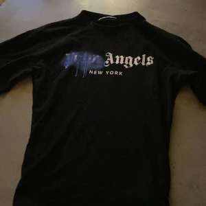 Svart t-shirt från Palm Angels - Säljer en svart t-shirt från Palm Angels med tryck i vitt och blått framtill där det står 'Angels New York'. Klassisk passform och rund halsringning. Perfekt för dig som gillar streetwear.