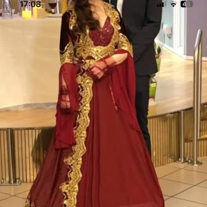 Henna klänning Vinröd långklänning med guldbroderi - Säljer en vinröd långklänning med vackra guldbroderier och vida ärmar. Klänningen har en elegant och traditionell look med detaljer i spets och guld längs hela framsidan och ärmarna. Perfekt för festliga tillfällen.