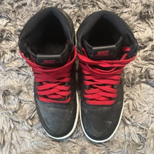 Svarta Nike Air Jordans med röda skosnören - Säljer ett par svarta Nike Air Jordans med röda skosnören och svart sula. Skorna har högt skaft och klassisk AirJordan -logga på sidan och plösen. Perfekta för dig som gillar streetstyle och vill sticka ut med Svart och rött.