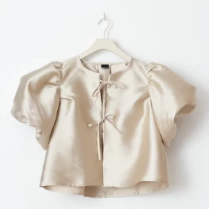 Beige satintopp med puffärmar och knyt - *oanvänd* Trendig beige topp i satin med stora puffärmar och två knytband framtill. Toppen har en lätt glansig yta och en rak, något croppad passform. Perfekt för dig som vill sticka ut med en unik och elegant look. Passar verkligen inför sommaren💞På bilden ser den silver ut på grund av skimret , men egentligen är den guld 🩷