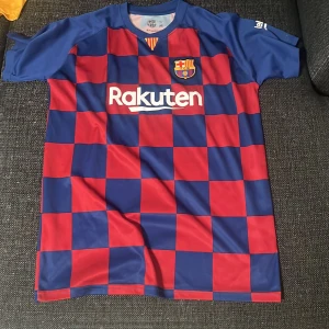 FC Barcelona Messi 10 fotbollströja - Säljer en FC Barcelona fotbollströja med Messi 10 på ryggen. Tröjan är kortärmad och har ett rödblått rutigt mönster med klubbmärke och Rakuten-tryck på framsidan. Materialet är lätt och andas, perfekt för fotboll eller support.