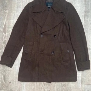 Brun kappa - Snygg brun kappa från Vero moda med dubbelknäppning och bred krage. Jackan har långa ärmar och två framfickor. Perfekt för höst och vinter med sin klassiska stil och varma material. Också perfekt till våren. Lite skador men inget som påverkar dess utseende på utsidan