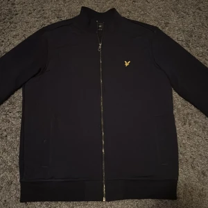 Svart jacka kan användas som kofta från Lyle & Scott - Snygg svart jacka från Lyle & Scott med dragkedja och ett broderat emblem på bröstet. Jackan har långa ärmar och en stilren design som passar perfekt för olika tillfällen.