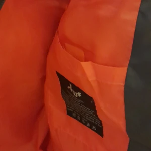 Grå dunväst från Stuff Clothing - Säljer en grå dunväst från Stuff Clothing med en kontrasterande orange insida. Västen har en dragkedja framtill och två fickor på sidorna. Perfekt för kyligare dagar. Oanvänd 