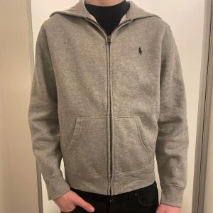 Grå hoodie från Ralph Lauren - Säljer en stilren grå hoodie från Ralph Lauren. Dragkedja och klassisk logga på bröstet, helt oanvänd. Storlek 14/16 år.