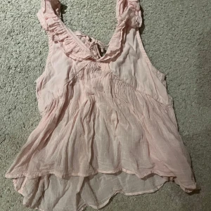 Ljusrosa blus från Gina Tricot - Säljer en söt ljusrosa blus från Gina Tricot med volangdetaljer och knytning i ryggen! Blusen är ärmlös och har en luftig passform, perfekt för varma dagar. Den har en romantisk och feminin stil som passar till många olika tillfällen!!🥰🥰😍😍❣️🤗