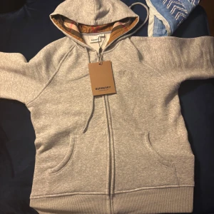 Grå hoodie från Burberry - Passar 160-165. Säljer en stilren grå hoodie från Burberry med dragkedja och fickor framtill. Den har en klassisk design med märkets ikoniska rutiga foder i huvan. 