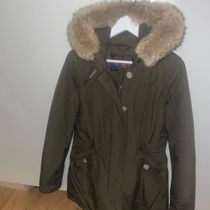 Grön parkajacka från Woolrich - Säljer en snygg grön parkajacka från Woolrich med pälskrage. Jackan har långa ärmar och stängs med både dragkedja och knappar. Perfekt för kyliga dagar! Jackan är i väldigt bra skick