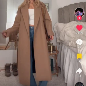 Beige kappa med bälte - Snygg beige kappa med knytbälte i midjan. Den har en klassisk design med långa ärmar och en slits baktill. Perfekt för att hålla stilen under kyligare dagar. Har hål i ena fickan