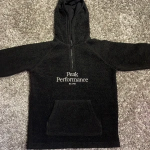 Svart fleece hoodie från Peak Performance - Säljer en svart fleece hoodie från Peak Performance med halv dragkedja och känguruficka. Perfekt för kyliga dagar med sin mjuka och varma känsla. Peak Performance-loggan är tryckt på framsidan. Passar både för vardag och utomhusaktiviteter.