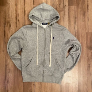Grå hoodie från Polo Ralph Lauren - Säljer en stilren grå hoodie från Polo Ralph Lauren med dragkedja och huva. Tröjan har en liten broderad logga på bröstet och praktiska fickor framtill. Perfekt för en avslappnad look.