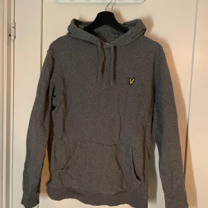 Grå hoodie från Lyle & Scott - Säljer en stilren grå hoodie från Lyle & Scott med deras ikoniska logga på bröstet. Tröjan har en klassisk känguruficka och justerbar huva med dragsko. Perfekt för en avslappnad look.