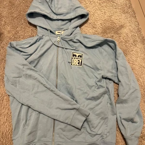 Ljusblå hoodie från Obey - Säljer en ljusblå hoodie från Obey med dragkedja och stort tryck på ryggen. 