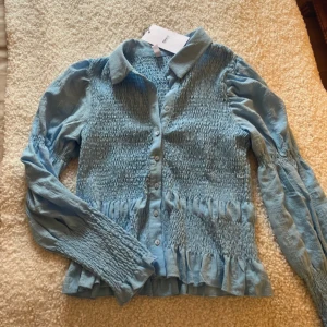 Blå blus från ONLY - Säljer en snygg blå blus från ONLY med smockdetaljer och knappar framtill. Blusen har långa ärmar och en klassisk krage. Perfekt för att ge en stilren look.