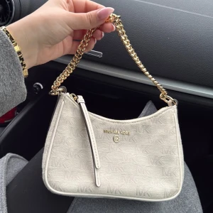 Beige axelväska från Michael Kors - Snygg beige axelväska från Michael Kors med guldkedja och diskret MK-mönster. Väskan har en dragkedja upptill och är perfekt för att bära dina viktigaste saker med stil. Den eleganta designen gör den till ett måste i garderoben.