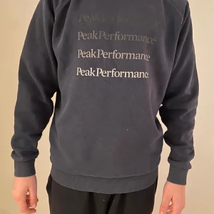 Mörkblå tröja från Peak Performance - Säljer en stilren mörkblå tröja från Peak Performance med deras logga tryckt flera gånger på framsidan. Tröjan har långa ärmar och ribbade muddar vid ärmslut och nederkant. Perfekt för en avslappnad stil.