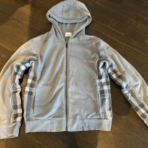 Blå hoodie från Burberry - Säljer en snygg blå hoodie från Burberry med klassiskt rutmönster längs ärmarna. Tröjan har dragkedja och huva, perfekt för en stilren look. Passar bra för både vardag och avslappnade tillfällen.