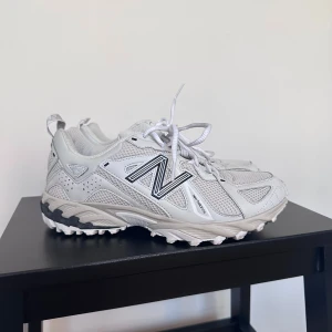 New Balance 610T - Säljer ett par vita New Balance sneakers med svart logga. Skorna har snörning och en robust sula som ger bra grepp. Perfekta för både vardag och träning. 
