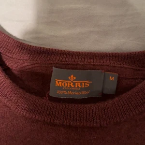 Vinröd merinoulltröja från Morris - Säljer en stilren vinröd tröja från Morris i 100% merinoull. Tröjan har långa ärmar och en liten broderad detalj på bröstet. Lite liten så skulle säga att den passar S bättre, Ett litet hål vid armen som inte synd vid användning