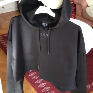 Svart croppad hoodie från SXS - Snygg svart croppad hoodie från SXS med en bekväm passform. Tillverkad i polyester och elastan för en mjuk känsla. Perfekt för en avslappnad stil med långa ärmar och en klassisk huva.