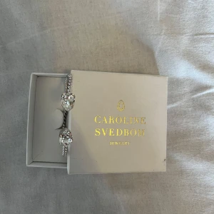 Silverfärgat armband från Caroline Svedbom - Elegant silverfärgat armband från Caroline Svedbom med en tvinnad design och stora, gnistrande stenar i ändarna. Sparsamt använd. 