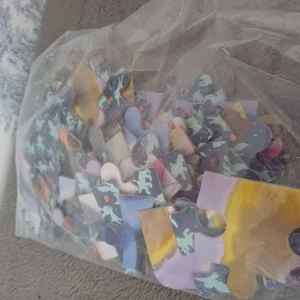 Pussel i plastpåse - Ett pussel med bitar i olika färger, förpackat i en genomskinlig plastpåse. Bitarna har varierande mönster och färger, inklusive blått, gult och lila. Perfekt för den som gillar att lägga pussel och vill ha en utmaning.