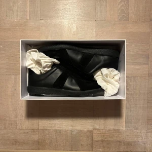 Maison margiela sneakers black - Tja, fräscha svarta sneakers från Maison Margiela med en stilren design. Skorna har en ovandel i skinn och mocka, samt en robust gummisula. Perfekta för dig som gillar en minimalistisk stil.